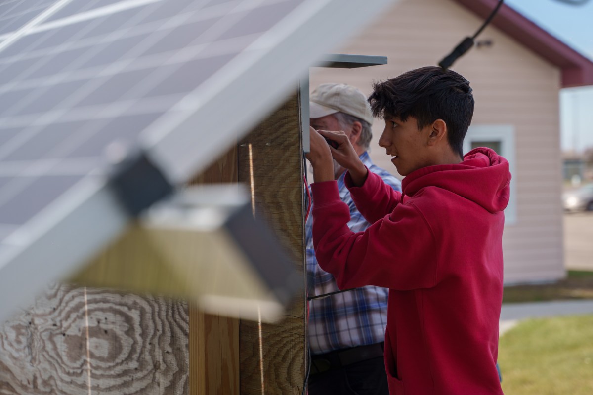 A young man in a red hoodie holds a tool and concentrates as he affixes a battery under a solar panel.