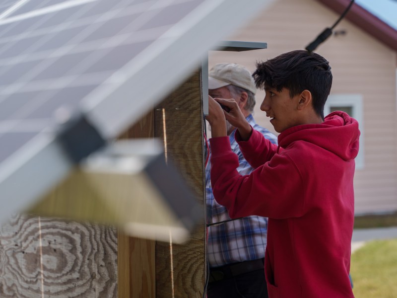 A young man in a red hoodie holds a tool and concentrates as he affixes a battery under a solar panel.