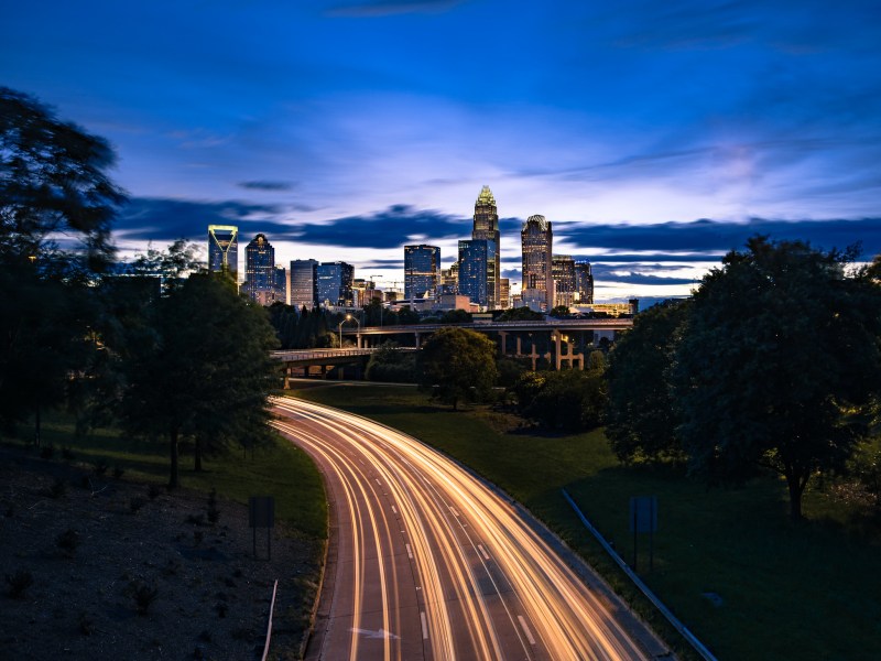 A long-exposure photo shows streaks of light from cars along a dark highway headed into the Charlotte skyline as the sun sets.