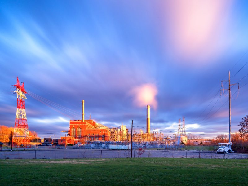 Power lines and a smoke stack are located around a large brick building, underneith a bright blue sky.