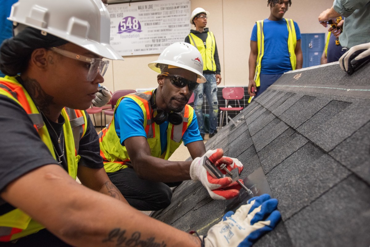 Students wearing safety gear practice installing brackets on a mockup of a pitched roof.