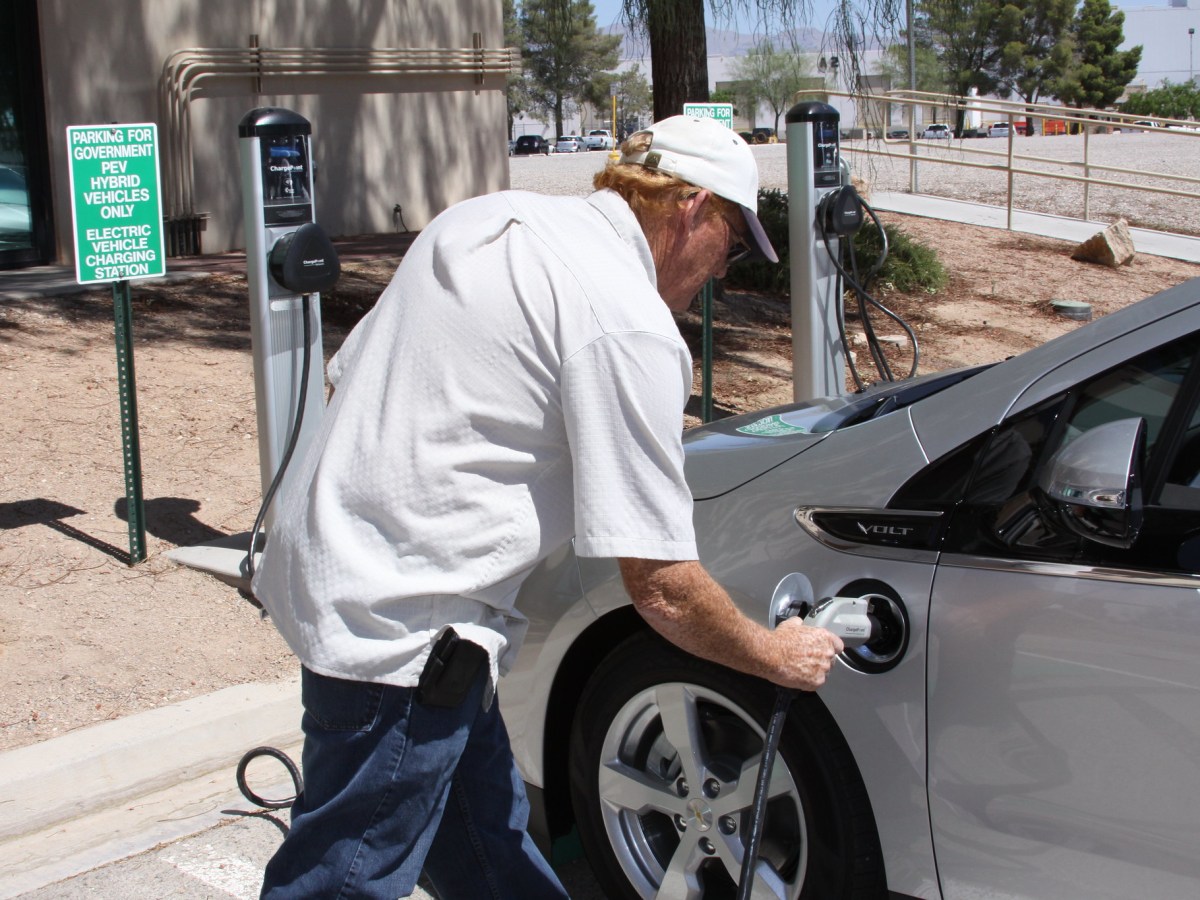 A man in a white shirt and baseball cap plugs in an electric vehicle in Las Vegas. The ground around him is a dustry red.