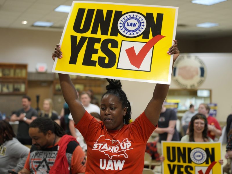 A woman in a red UAW t-shirt holds a yellow sign reading "Union Yes" over her head