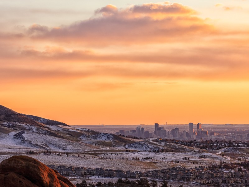 A distant view of Denver, Colorado showing suburban homes encroaching on the Rocky Mountain foothills with a yellow sunset in the background.