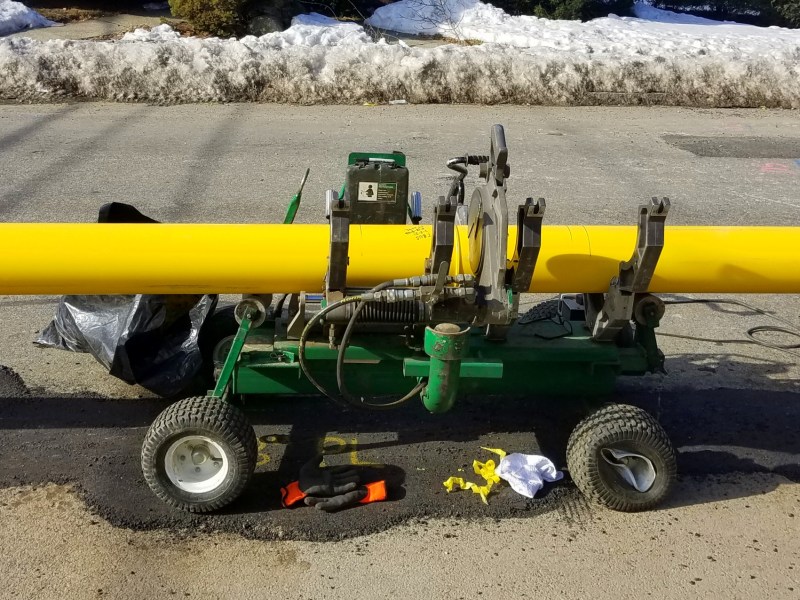 A green cart-like machine holds a yellow natural gas pipeline on the street.