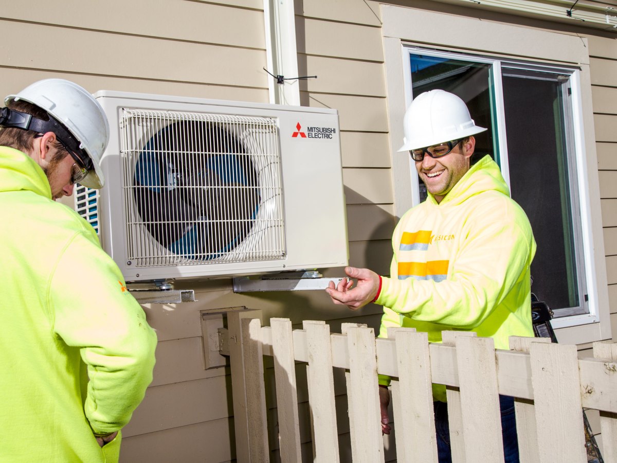 Jovial workers in hard hats installing a heat pump on the side of a house.