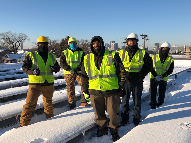 A group of people in reflective vests stand on a roof surrounded by snow-covered solar panels.