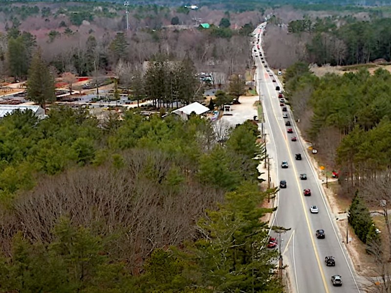 A video still showing heavy traffic on a two-lane highway through a wooded area of Maine that also features homes and commercial development.