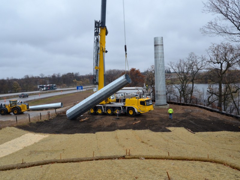 A heavy construction crane lifts a segment of a transmission tower into place along a rural expressway.