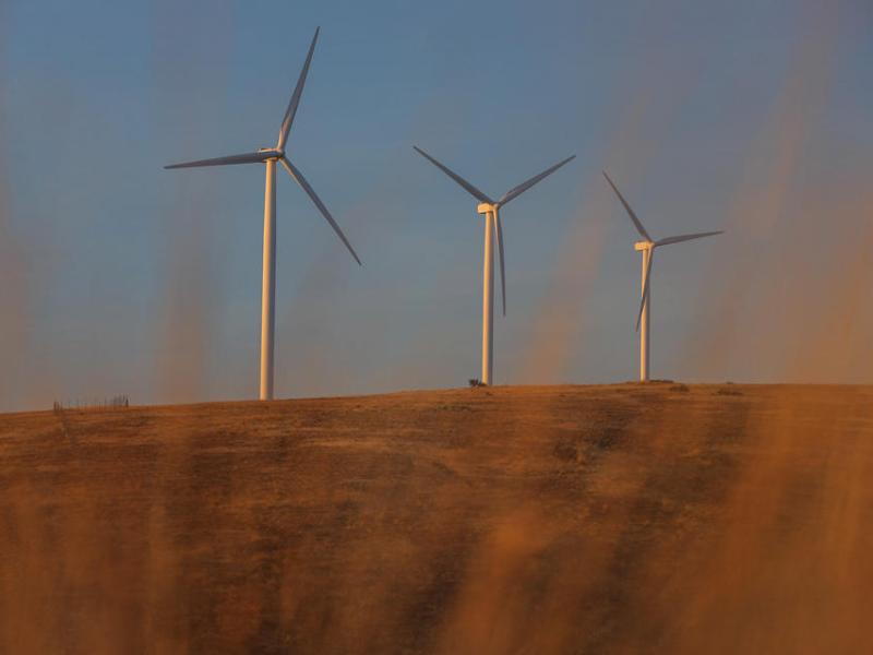 Three tall wind turbines sit on a brownish-orange hill against a blue sky.