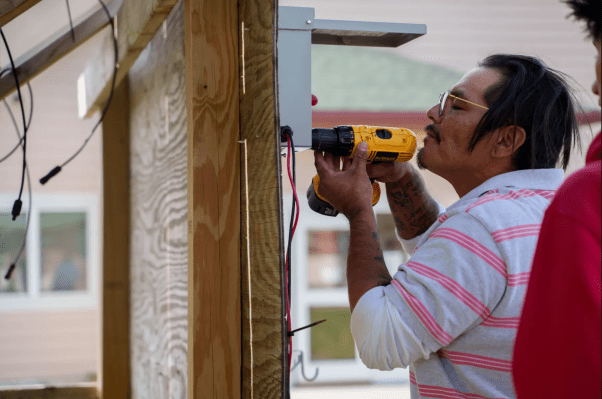 A man in a striped T-shirt holds a drill as he affixes a battery under a solar panel.