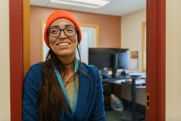 A woman wearing an orange beanie, glasses, a lanyard and a blue blazer stands smiling in a doorway